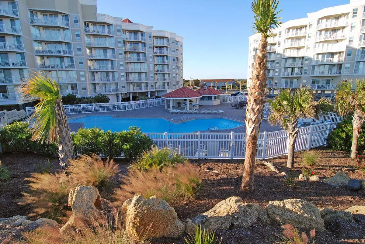 A vibrant outdoor swimming pool surrounded by tropical landscaping, with a cabana featuring a red roof and residential buildings in the background. The pool area invites relaxation, socializing, and sunbathing, while palm trees and lush greenery contribute to the resort-like ambiance.