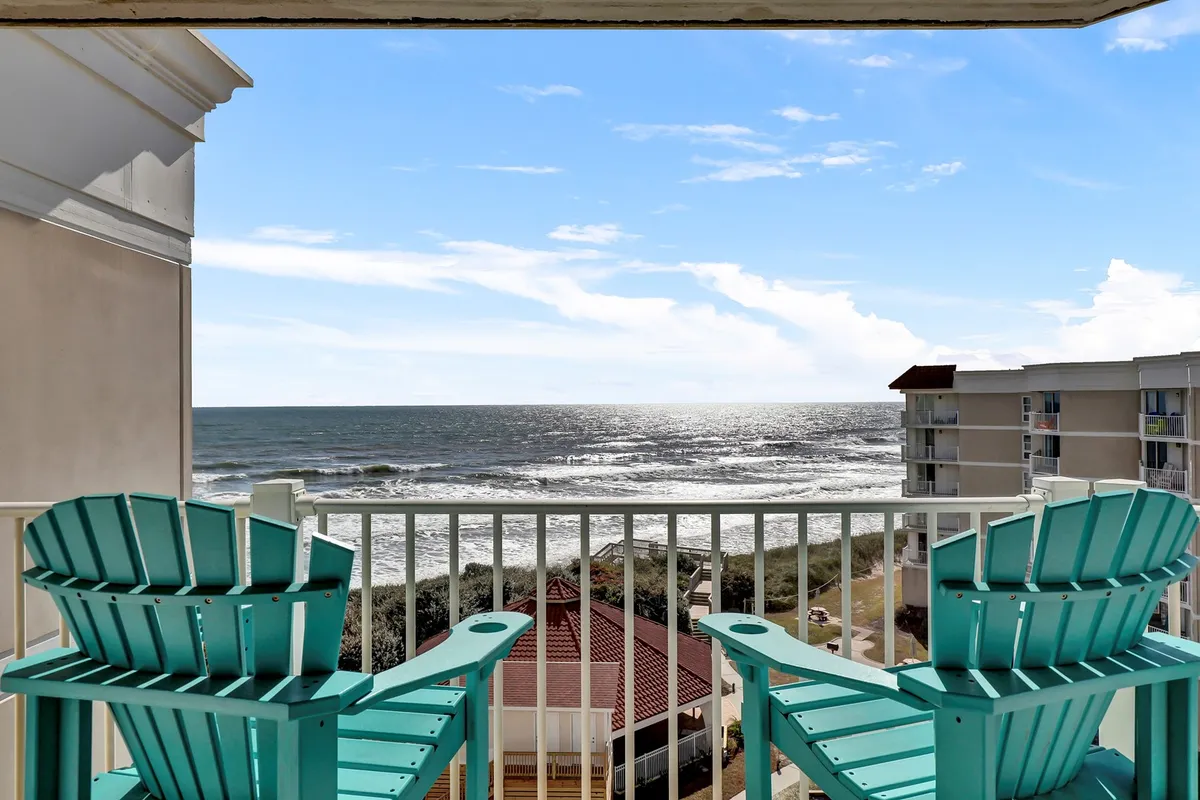 The image showcases two inviting turquoise Adirondack chairs set on a balcony overlooking the sparkling Atlantic Ocean. Sunlight dances on the water, suggesting a tranquil atmosphere perfect for sunrise coffees, sunset toasts, or quiet afternoons with a book. The view highlights the condo’s prime beachfront location, offering a front-row seat to North Topsail Beach’s natural beauty.