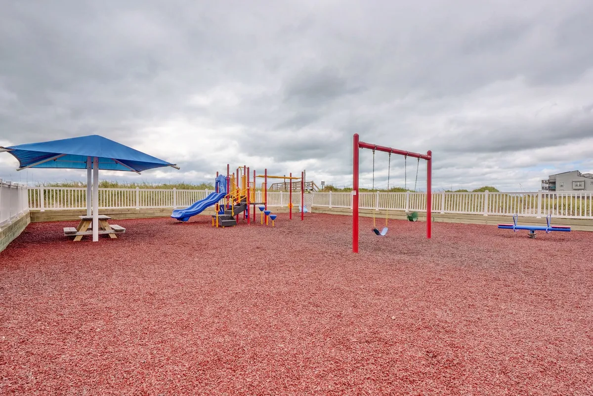 A colorful playground equipped with swings, slides, a sandbox, and a picnic table, set amidst trees and open space. Children are seen playing energetically, while adults enjoy relaxed moments nearby, creating a joyful family-friendly scene.