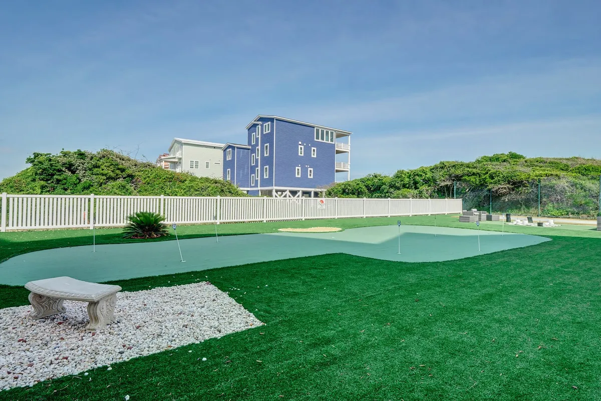 A lush green putting green bordered by white stones, with landscaped greenery and a blue house in the background. The space is open and bathed in sunlight, ideal for casual outdoor play and relaxation.