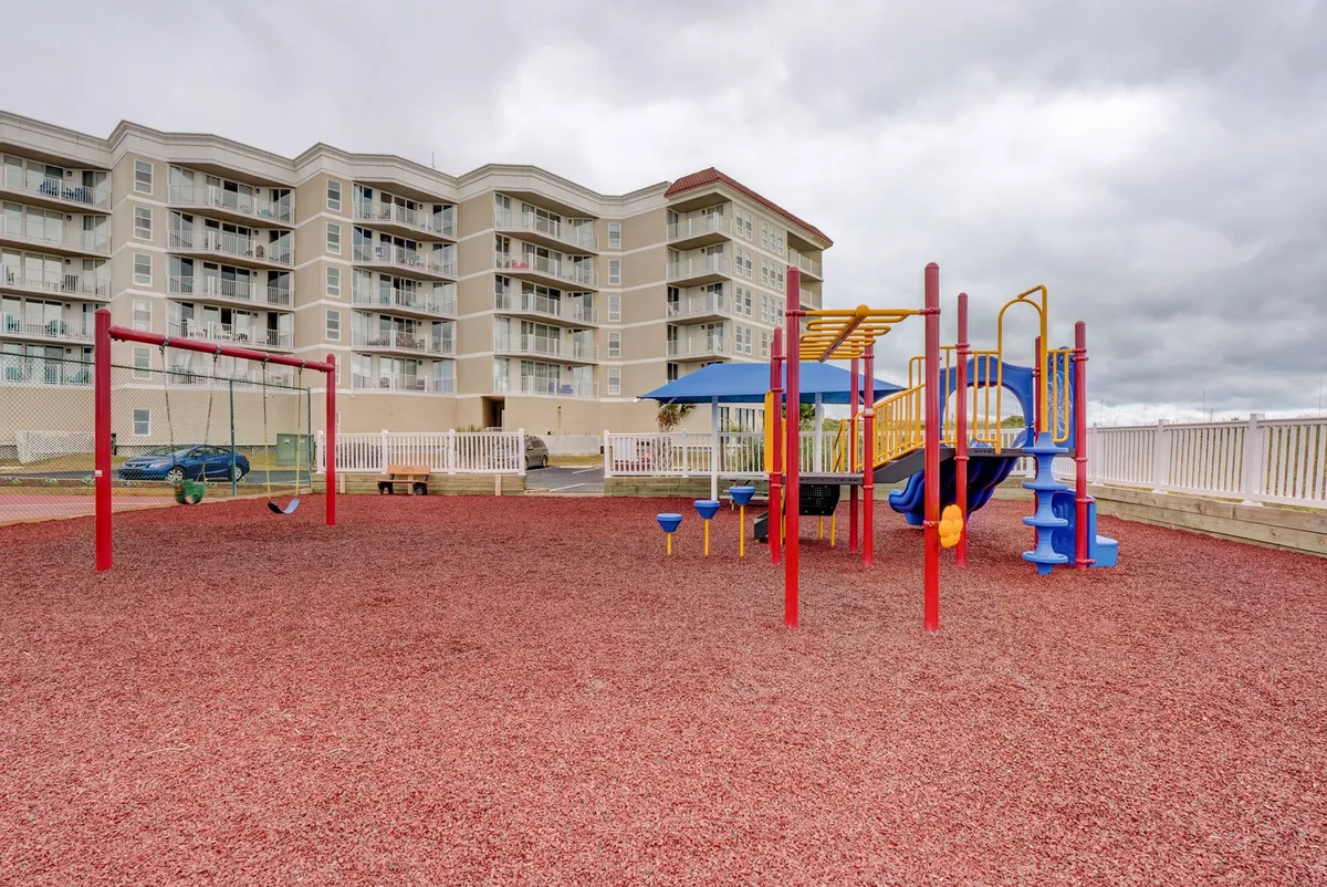 A modern, colorful playground with swings and play structures set on a red rubber surface, designed for children’s enjoyment. Families can be seen gathering around, with kids actively playing and adults watching nearby, evoking a sense of community and safety.