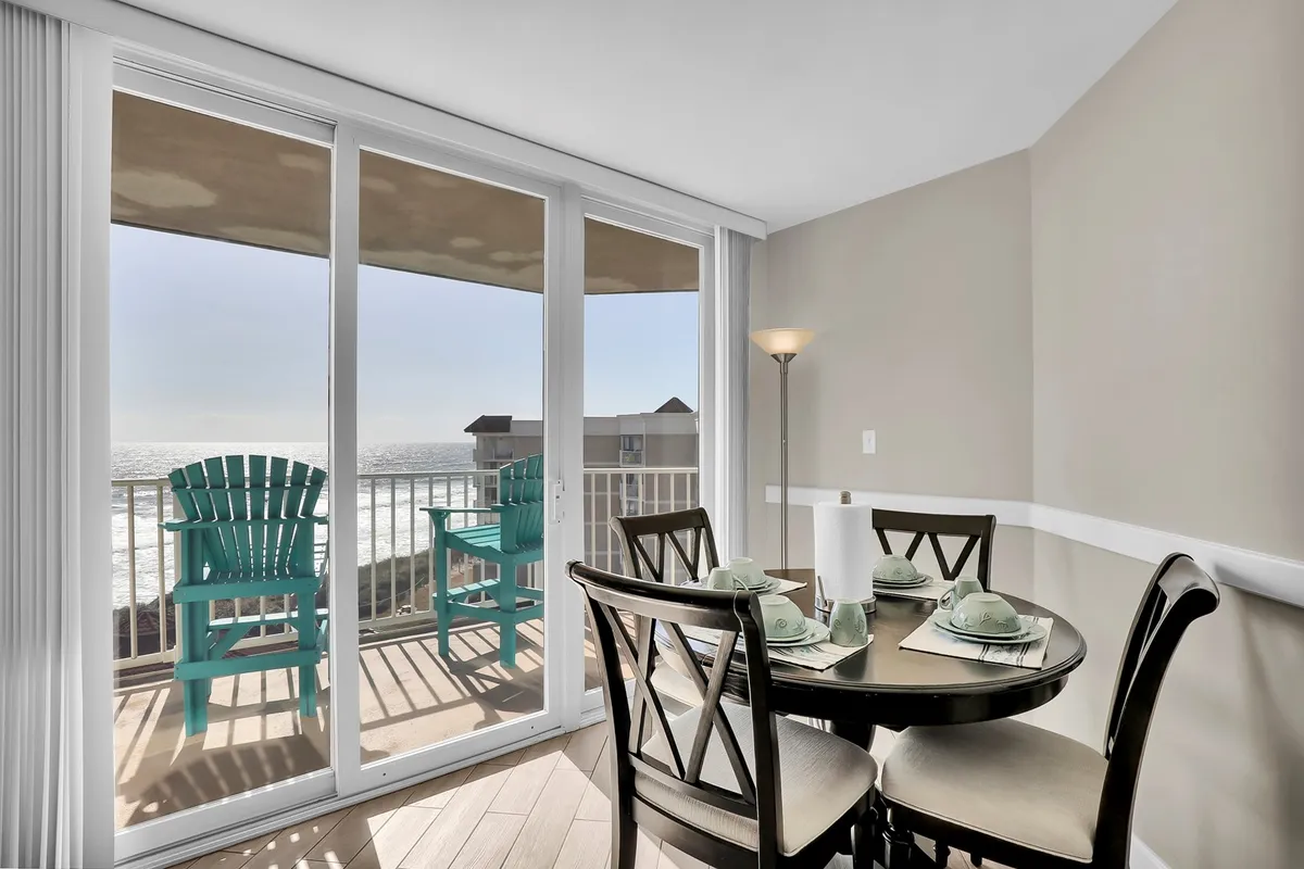 A cheerful dining area with a table set near expansive windows, revealing adirondack chairs and a striking ocean view beyond. The scene radiates brightness, encouraging shared meals, conversations, and quiet moments with the sea as your backdrop.