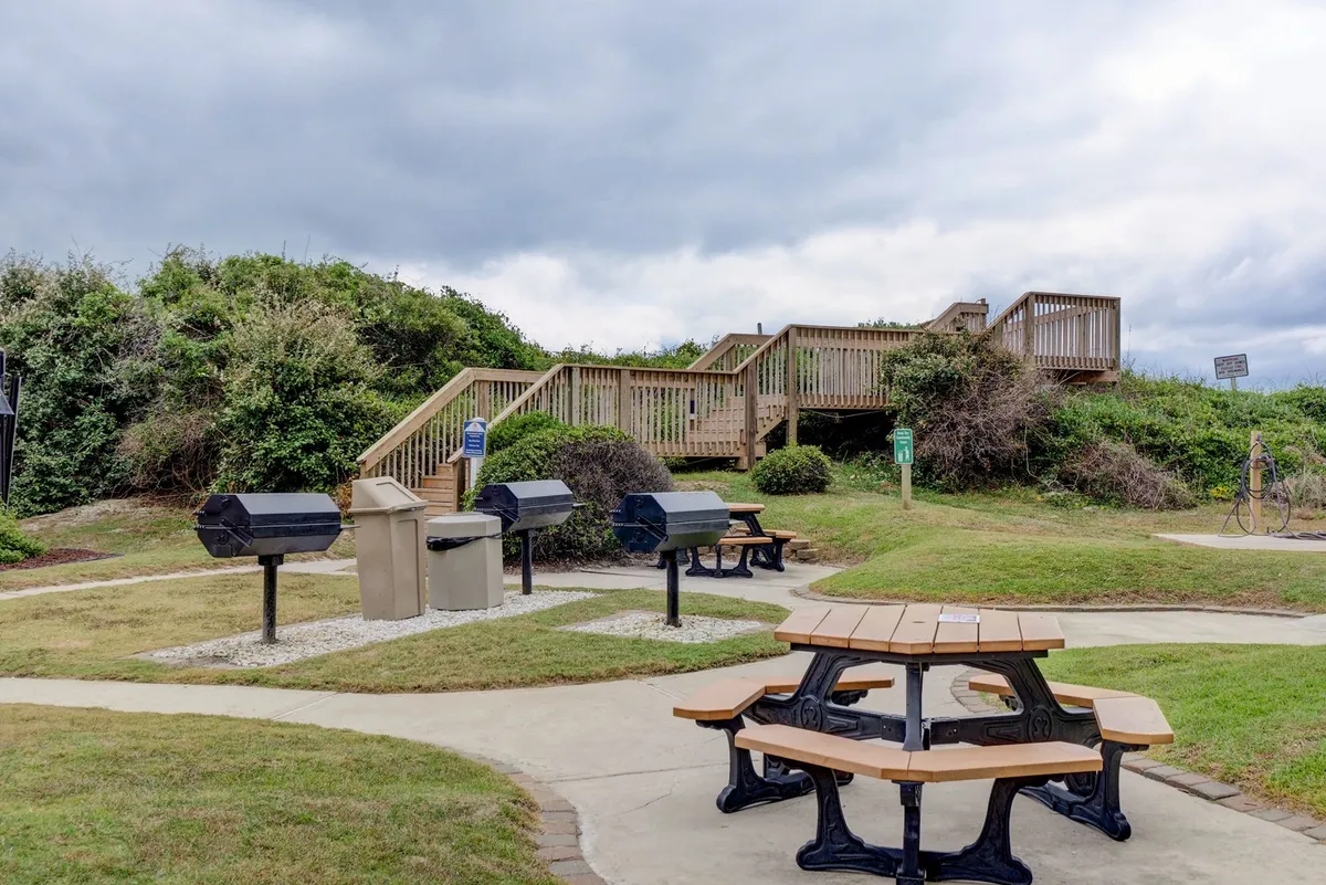 A welcoming outdoor area with picnic tables, barbecue grills, and a stairway leading up to the property. The space is designed for group gatherings, casual meals, and enjoying the fresh air, with all the essentials for a classic cookout.