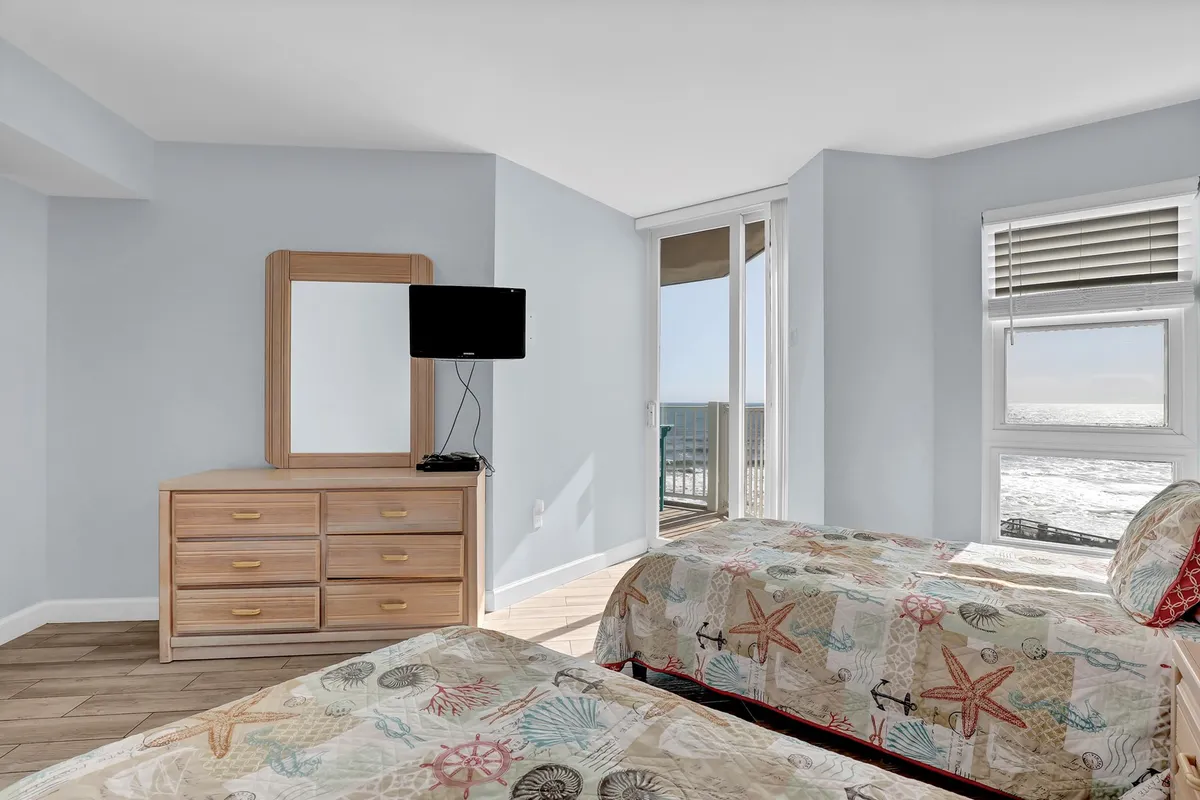A bright, tranquil bedroom with two beds adorned in vibrant bedding, a dressing table with a mirror, and a large window offering sweeping ocean views. The room is bathed in natural light, creating a soothing and refreshing atmosphere ideal for relaxation after a day at the beach.