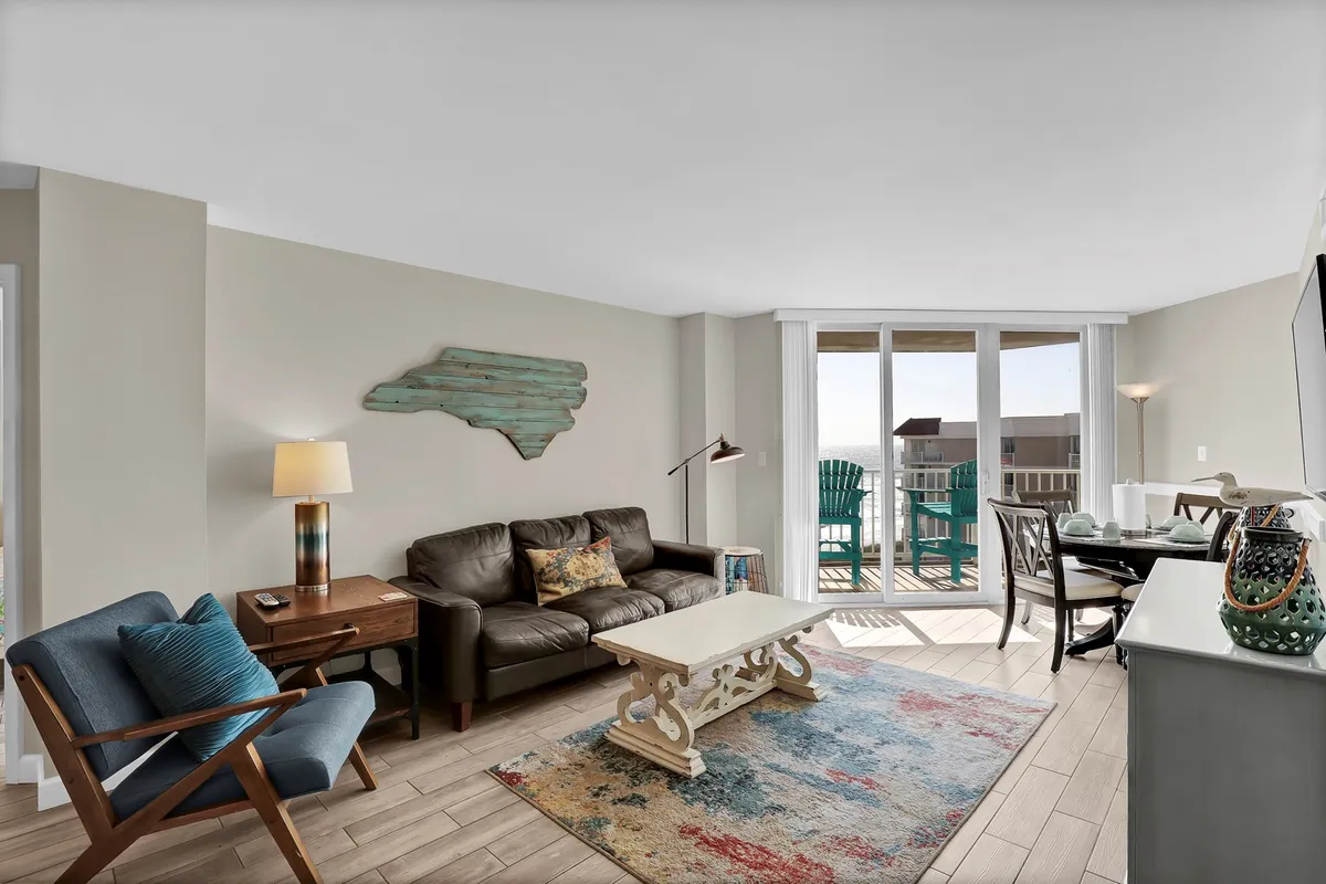 A cozy living room with a leather sofa, decorative rug, and wooden coffee table, all oriented towards a large window that frames a stunning ocean view. The space is bathed in natural light and invites relaxation and connection, with the sea visible right from your seat.